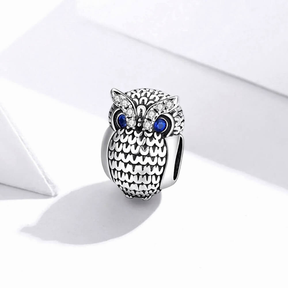 Sterling silver owl charm with textured feather design, blue eyes, and clear gemstones.