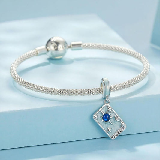 A silver-toned braided bracelet with a charm shaped like a tarot card featuring a blue gemstone and the word 'THE STAR', displayed on a light blue block.