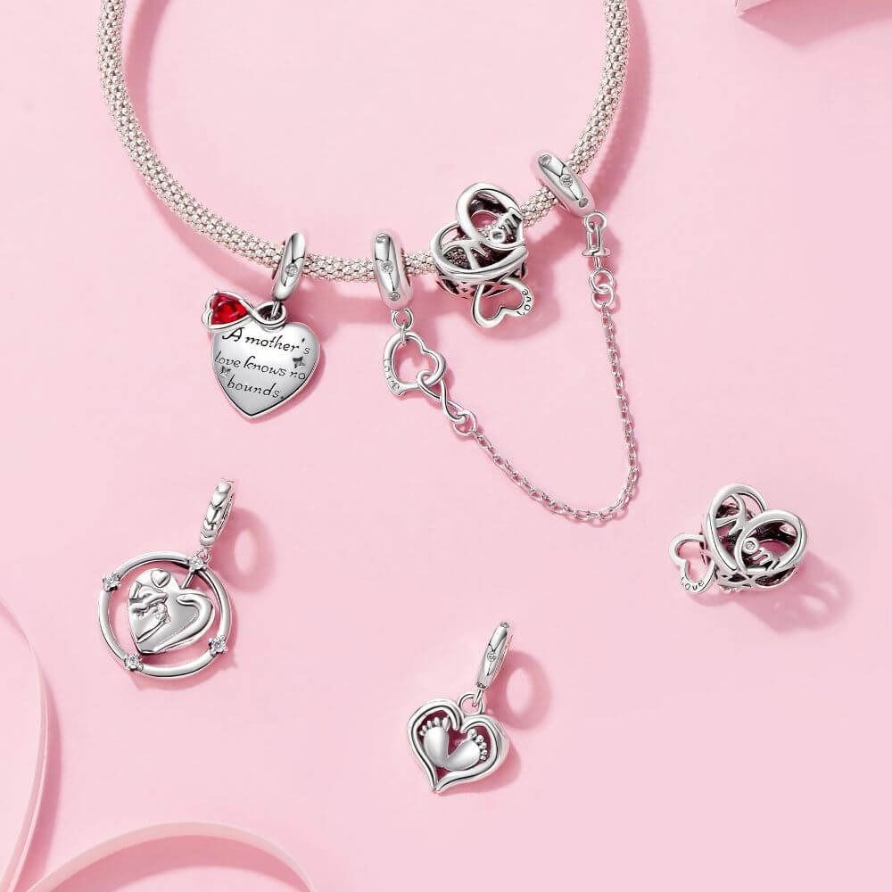 Silver heart-themed charm bracelet and matching pendant charms arranged on a soft pink background. The bracelet has heart-shaped charms, including one engraved with 'A mother is my best friend' with a small red heart accent, and other heart motifs.