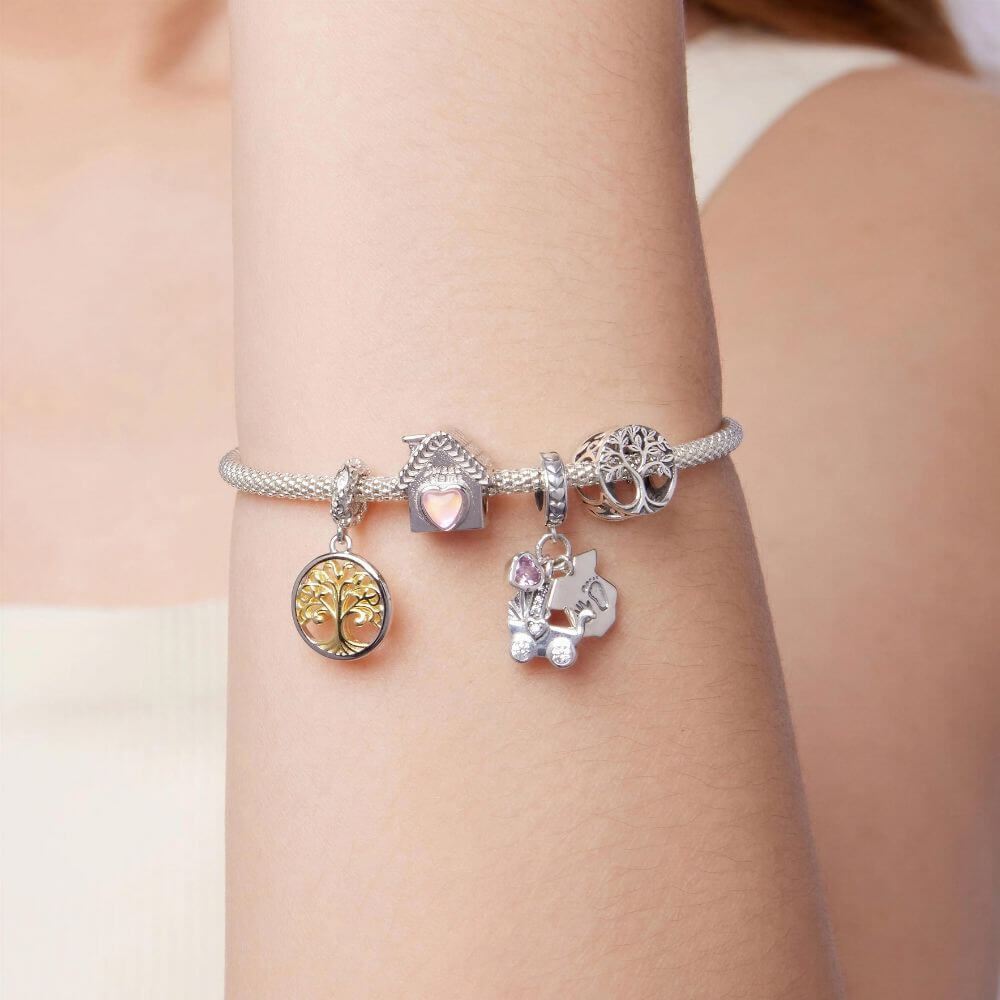 A close-up of a silver charm bracelet worn on a person's wrist. The bracelet features multiple charms, including a gold tree of life, a silver house with a pink heart, a silver owl, and a silver baby carriage with a small pink gem.