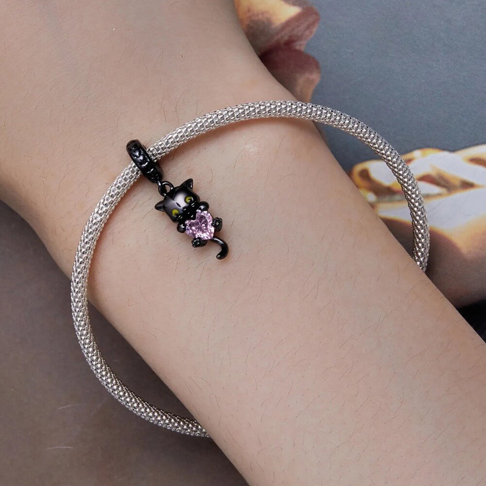 Black-plated silver cat charm holding pink heart-shaped gemstone with green eyes.