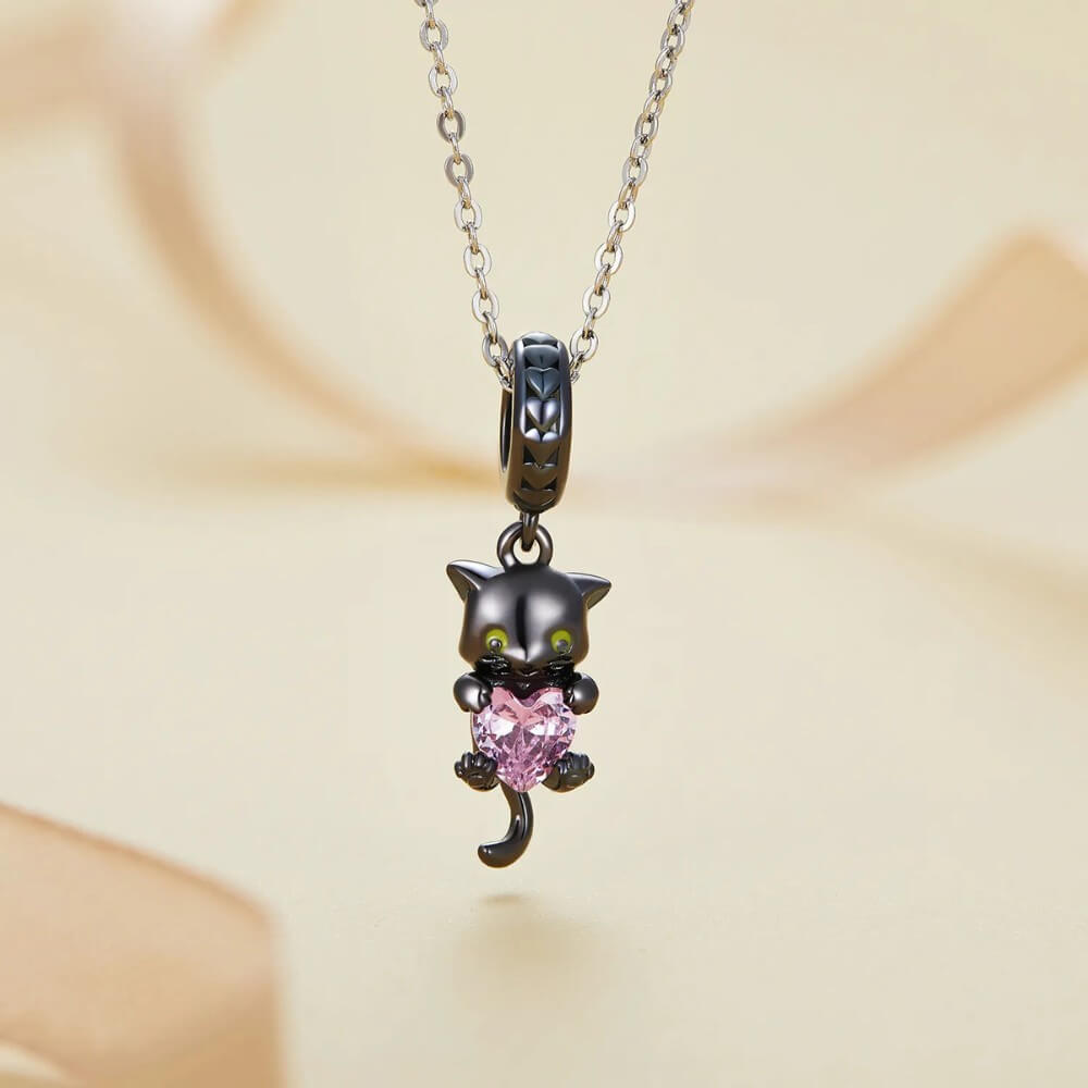 Black-plated silver cat charm holding pink heart-shaped gemstone with green eyes.