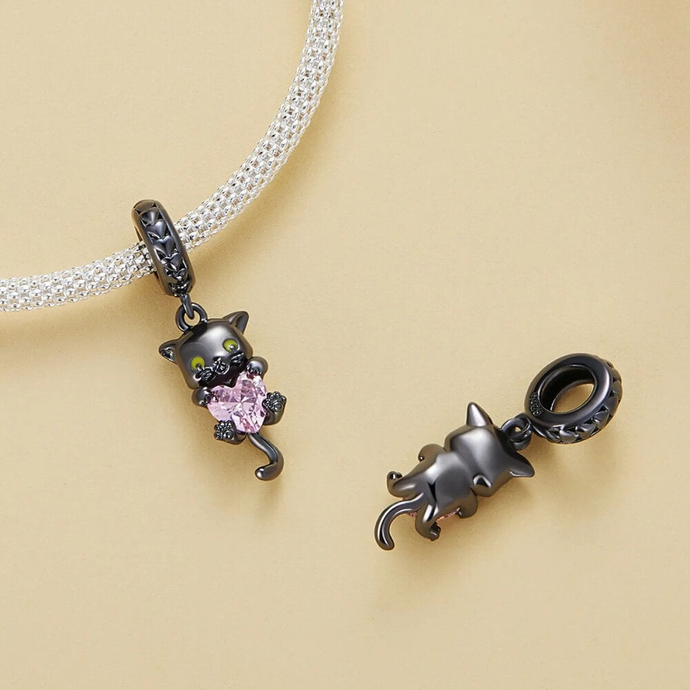 Black-plated silver cat charm holding pink heart-shaped gemstone with green eyes.