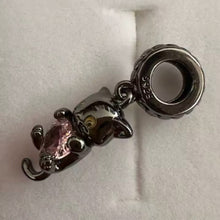 Black-plated silver cat charm holding pink heart-shaped gemstone with green eyes.