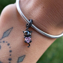 Black-plated silver cat charm holding pink heart-shaped gemstone with green eyes.