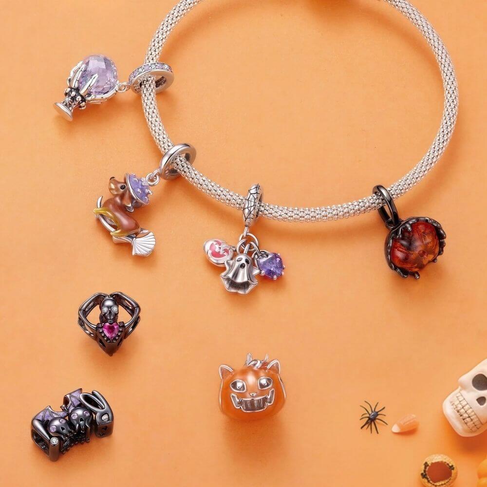 A silver charm bracelet displayed on an orange background, featuring multiple Halloween-themed charms including a cat head, skull, spider, and purple crystal accents. Several additional charms are arranged beside the bracelet.