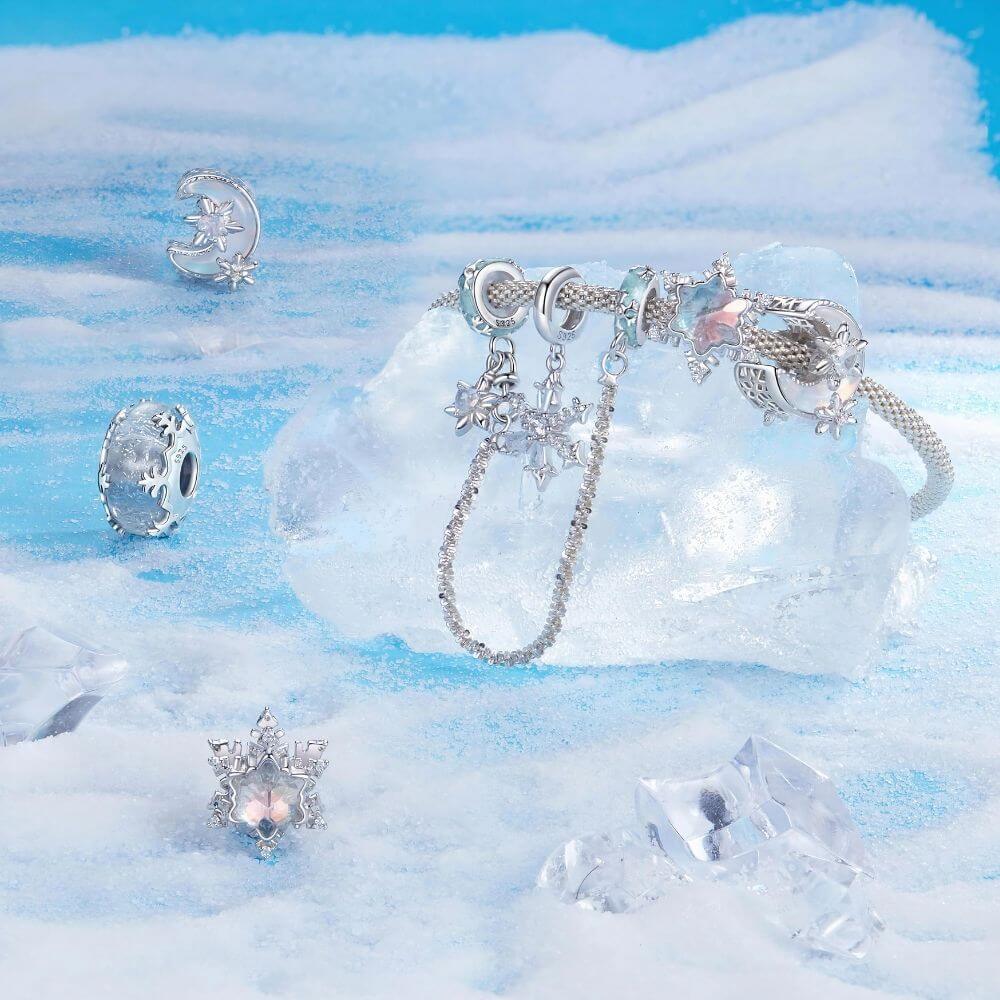 A silver-toned bracelet with multiple dangling charms, displayed on a translucent ice block with matching icy jewelry pieces scattered around it on a blue and white textured surface.