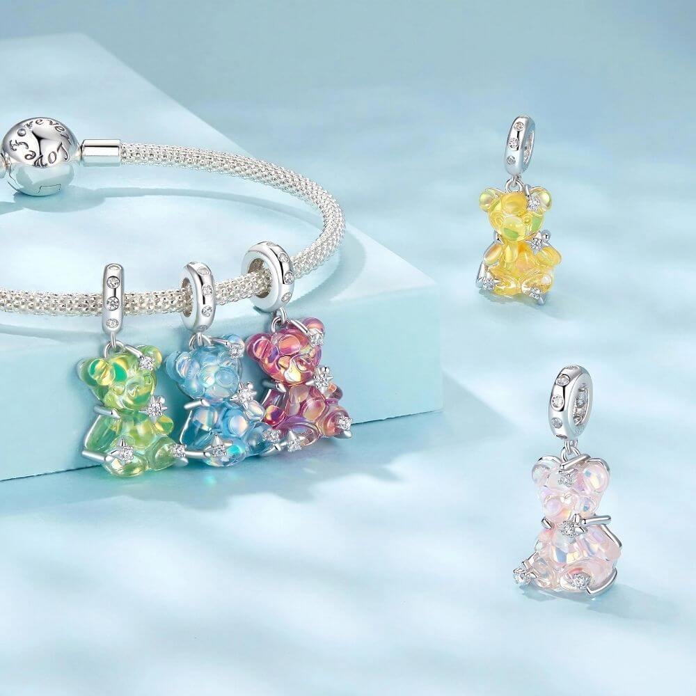 Silver charm bracelets featuring colorful iridescent bear charms, displayed on a light blue background with one bracelet laid flat and three charms shown individually.