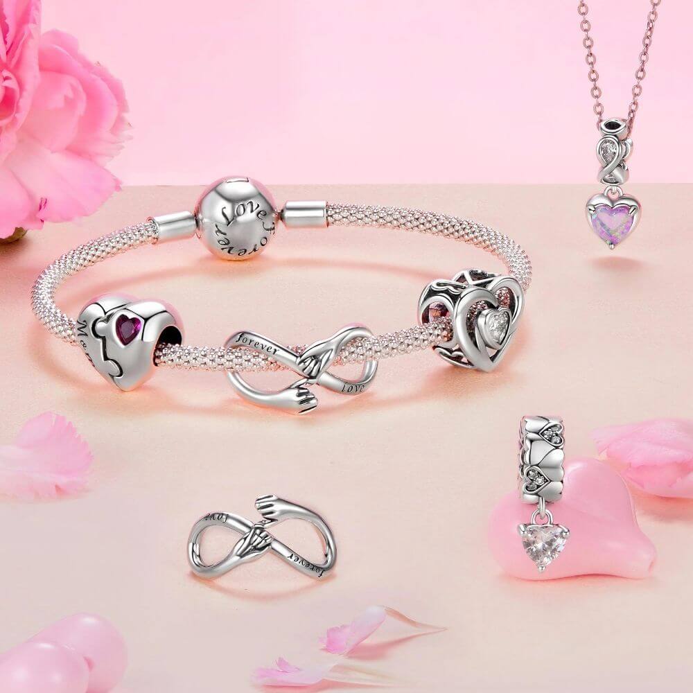Silver-toned jewelry set featuring a bracelet, necklace, ring, and charm pieces with heart and infinity motifs, arranged on a soft pink background with decorative rose petals.