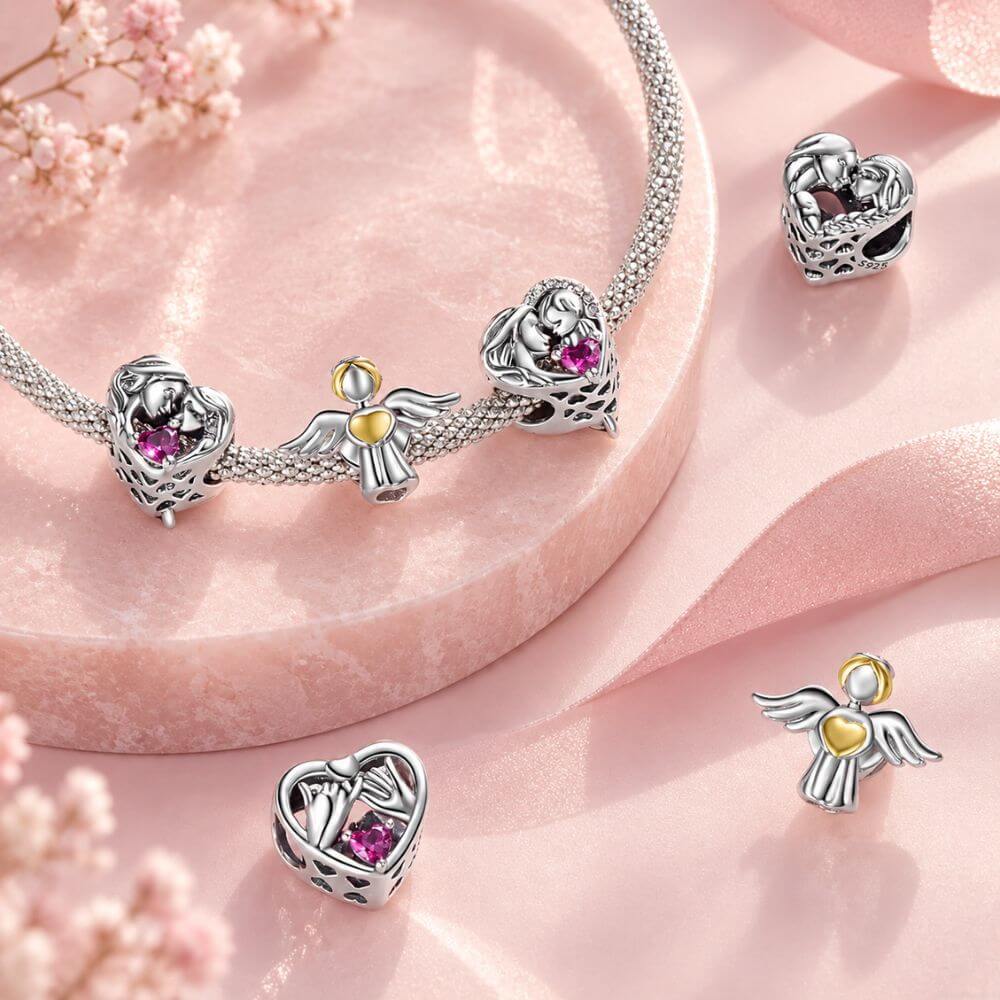 Silver-toned beaded jewelry charms displayed on a pink satin surface, including heart-shaped nesting birds with pink gemstones and angel figures with yellow and pink accents.