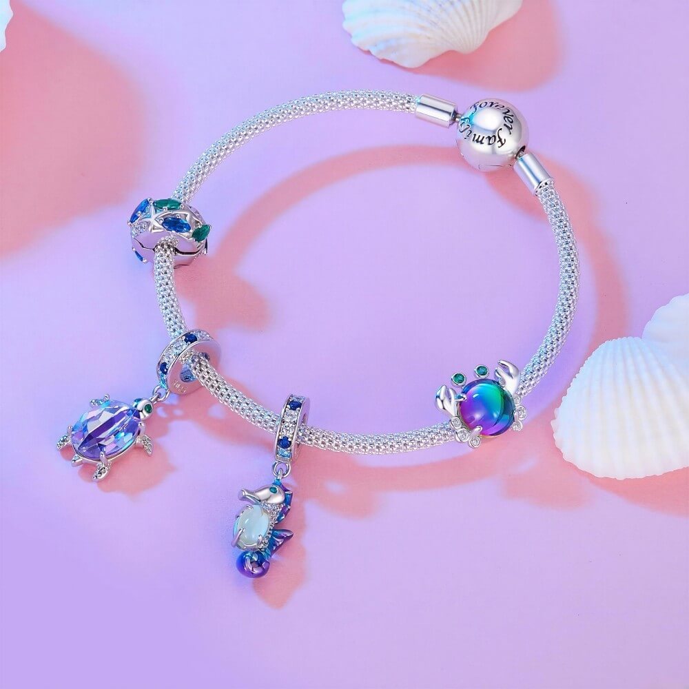 A silver-toned charm bracelet styled with sea turtle charms and iridescent blue-purple crystals, displayed on a pink background with white seashells.