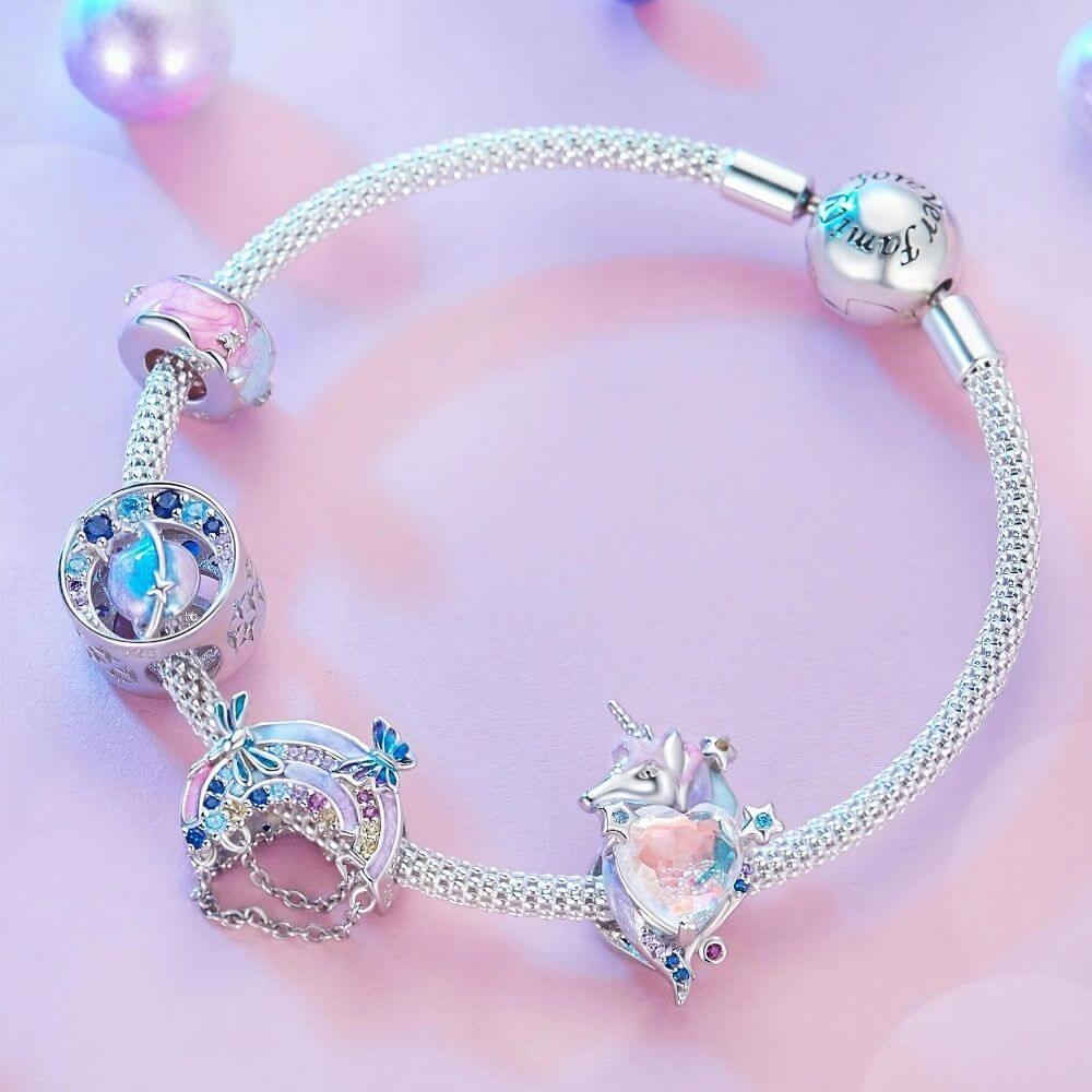 A silver chain bracelet with multiple charms, including a unicorn heart charm, a rainbow charm, and a blue gem charm, displayed on a soft pink background.