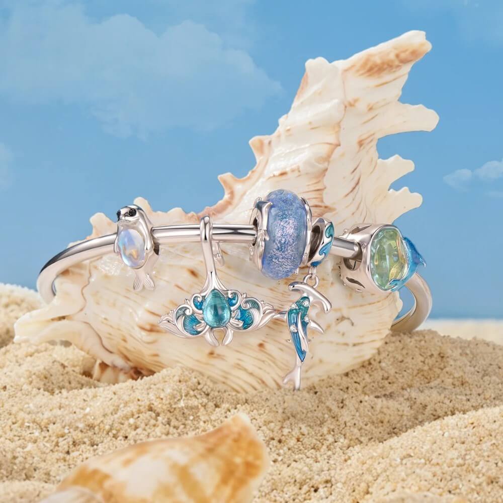 A silver bangle bracelet with blue and green bead details, displayed on a large seashell over sand with a blue sky background.