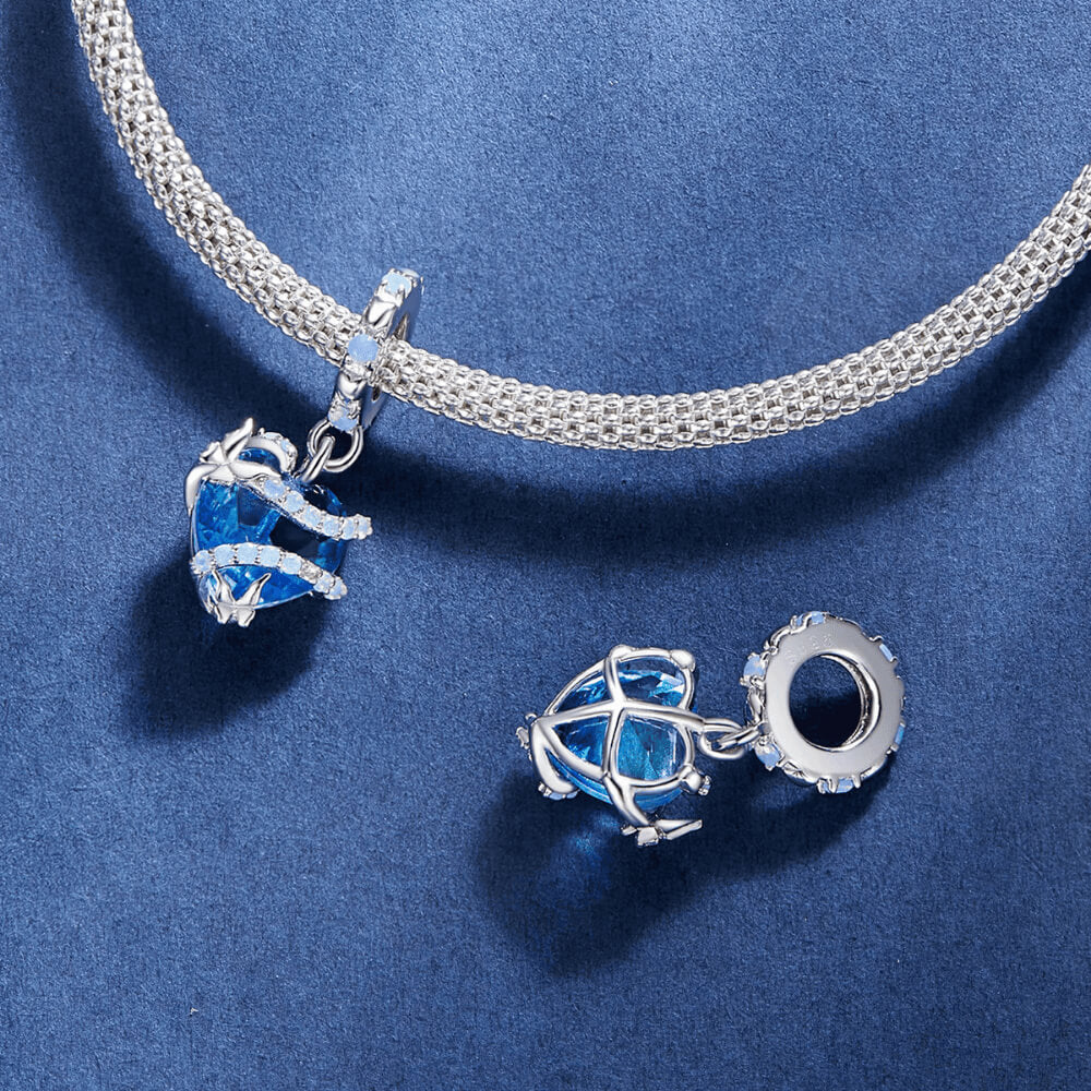 Sterling silver charm with a bright blue faceted heart crystal wrapped in silver vine-like details and small round light-blue stones on the bail 