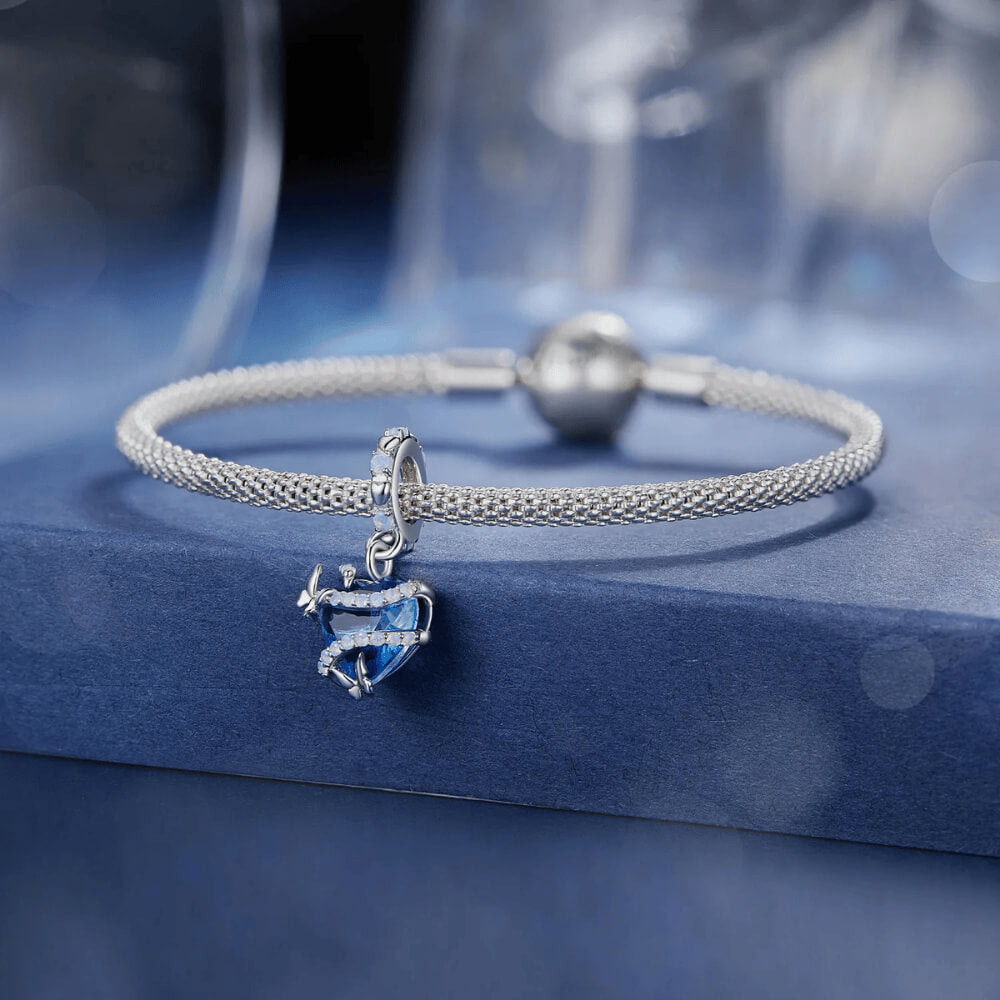 Sterling silver charm with a bright blue faceted heart crystal wrapped in silver vine-like details and small round light-blue stones on the bail 