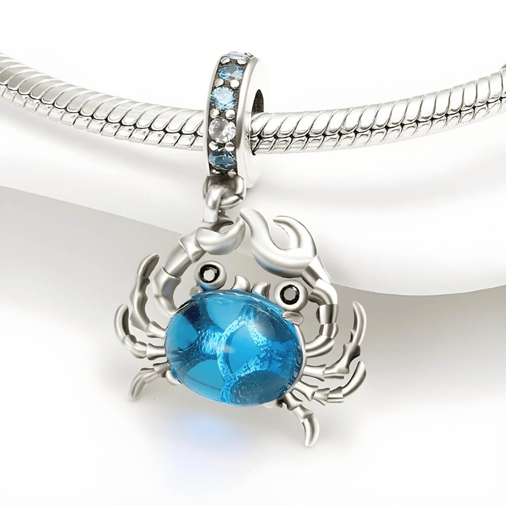 Sterling silver crab charm with a large blue oval stone body, black stone eyes, and a bail decorated with light-blue crystals 