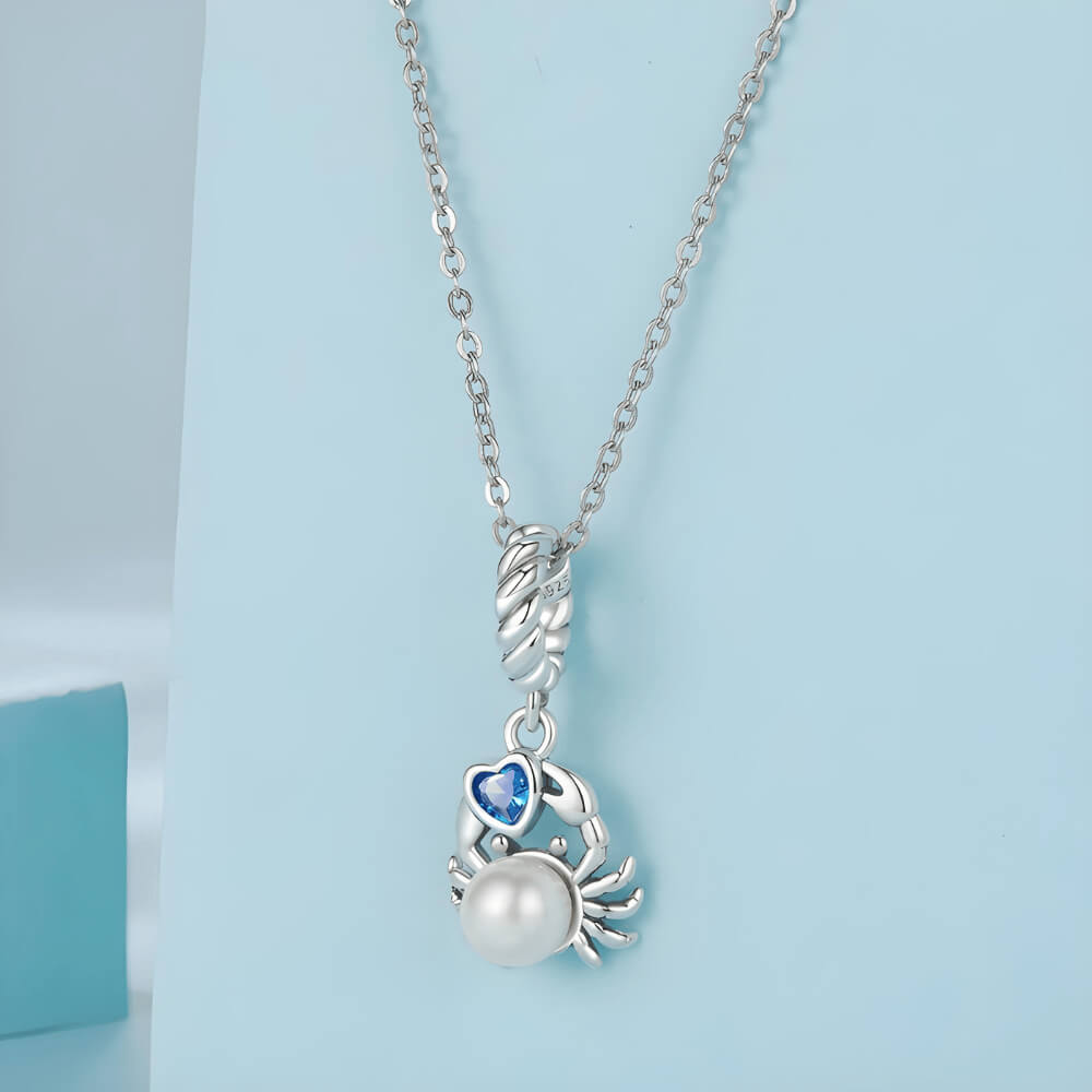 Sterling silver crab charm with a large white pearl-like body and a blue heart-shaped stone, on a twisted silver bail