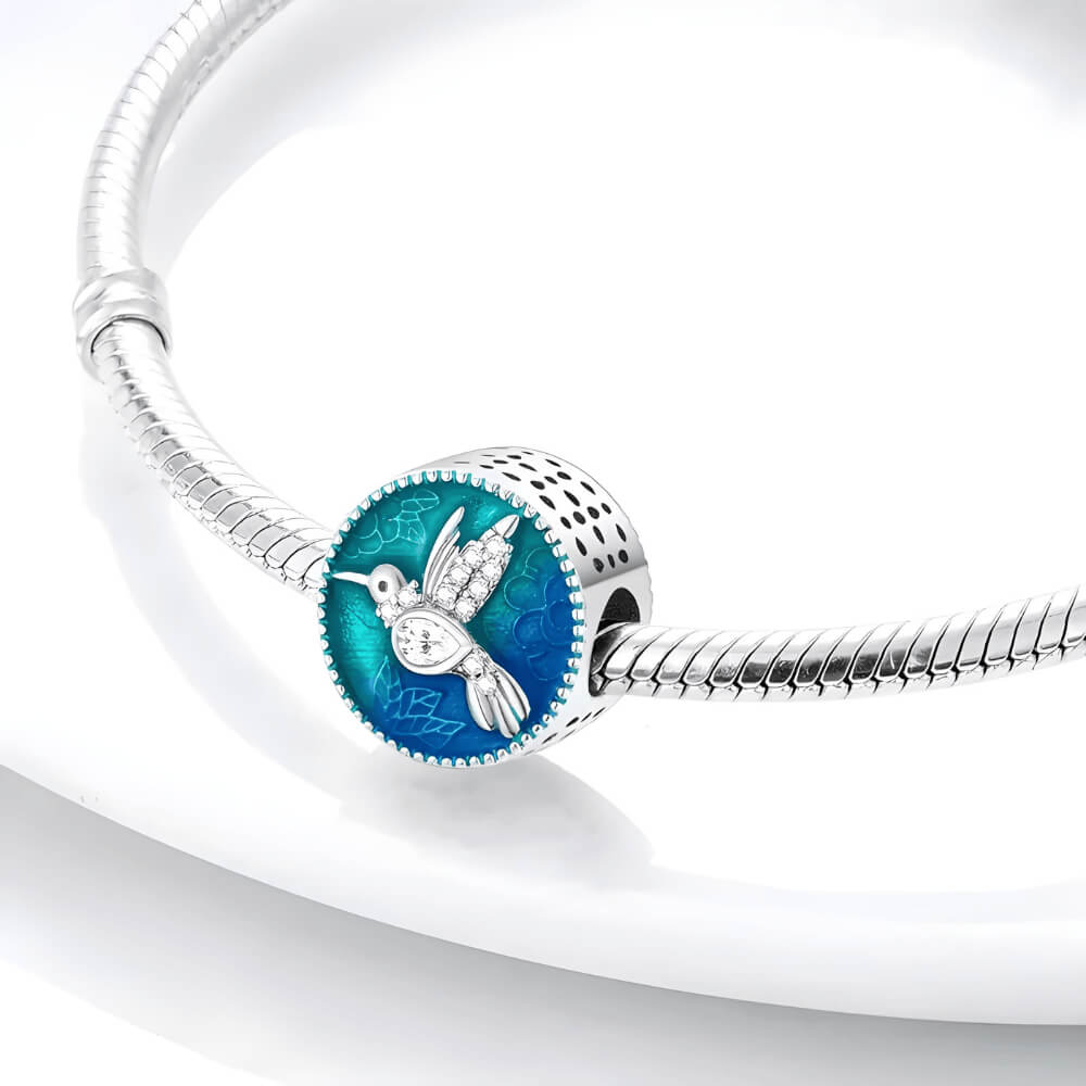 Sterling silver round charm with a crystal-studded hummingbird over a blue and teal enamel background with floral engravings