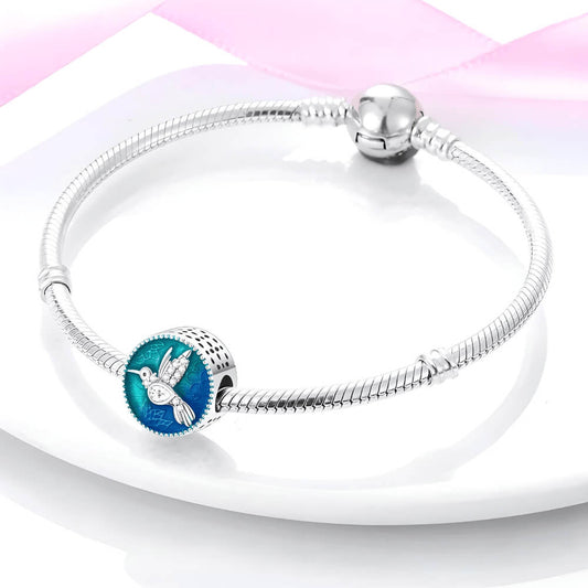Sterling silver round charm with a crystal-studded hummingbird over a blue and teal enamel background with floral engravings 