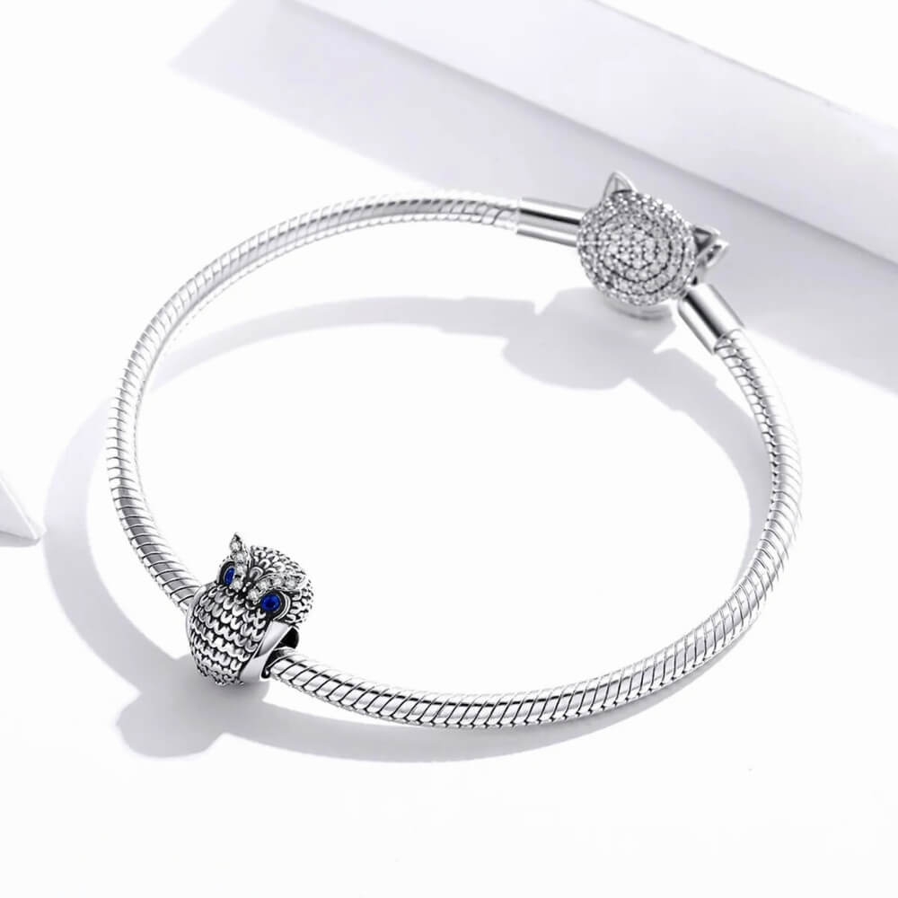 Sterling silver owl charm with textured feather design, blue eyes, and clear gemstones.