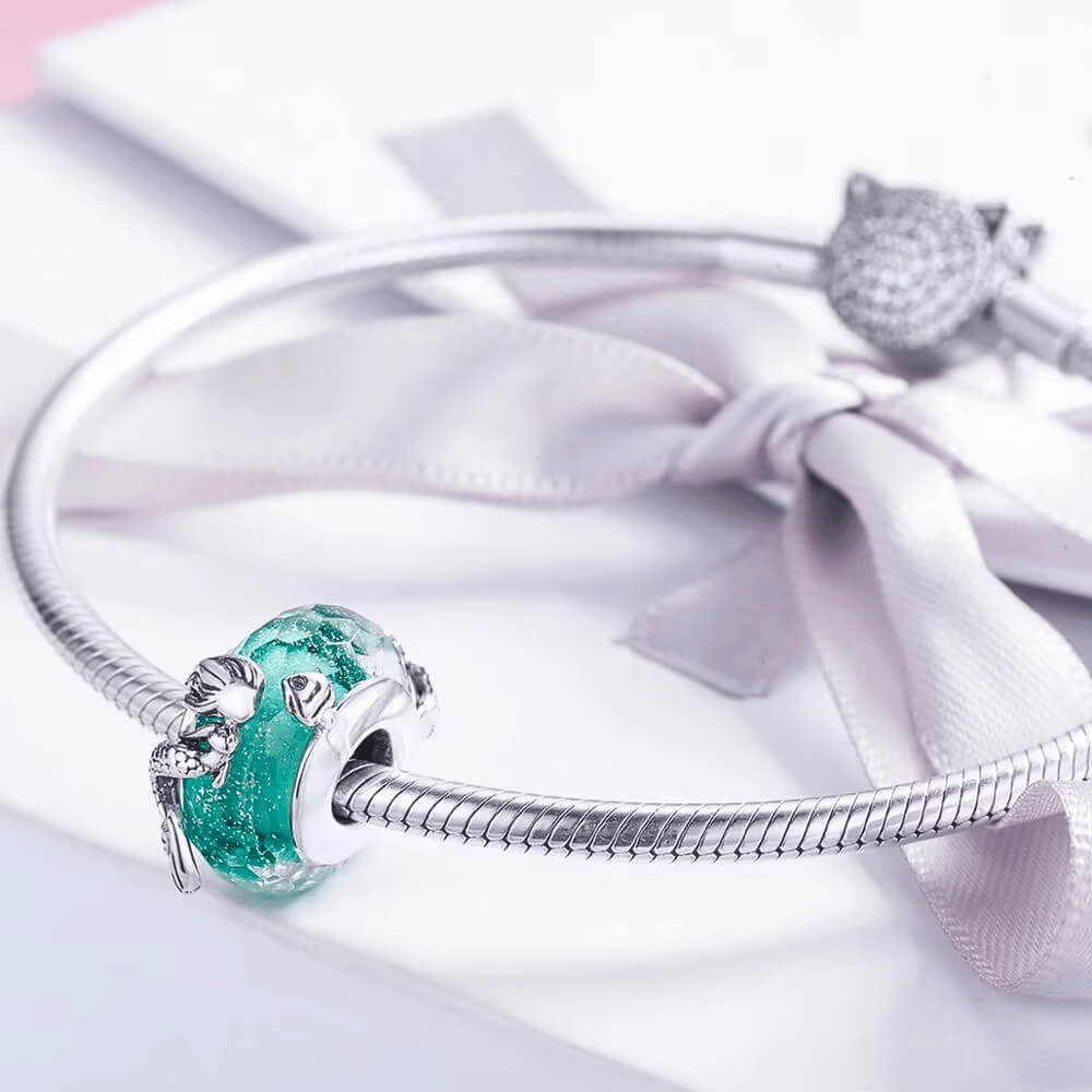 Sterling silver mermaid charm wrapped around teal faceted glass bead with textured details.