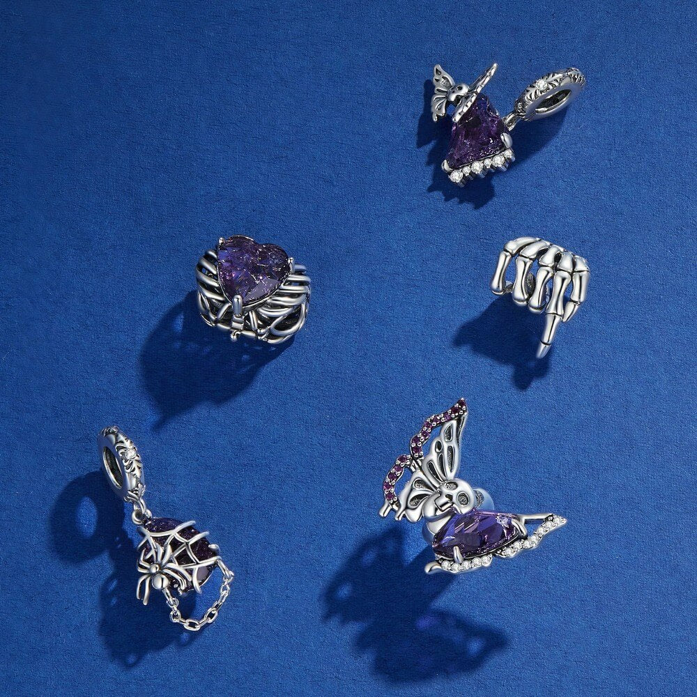 Five silver gothic-themed jewelry charms with purple gemstone accents, including a butterfly, skeleton hand, spiderweb, and heart motifs, arranged on a textured blue background.