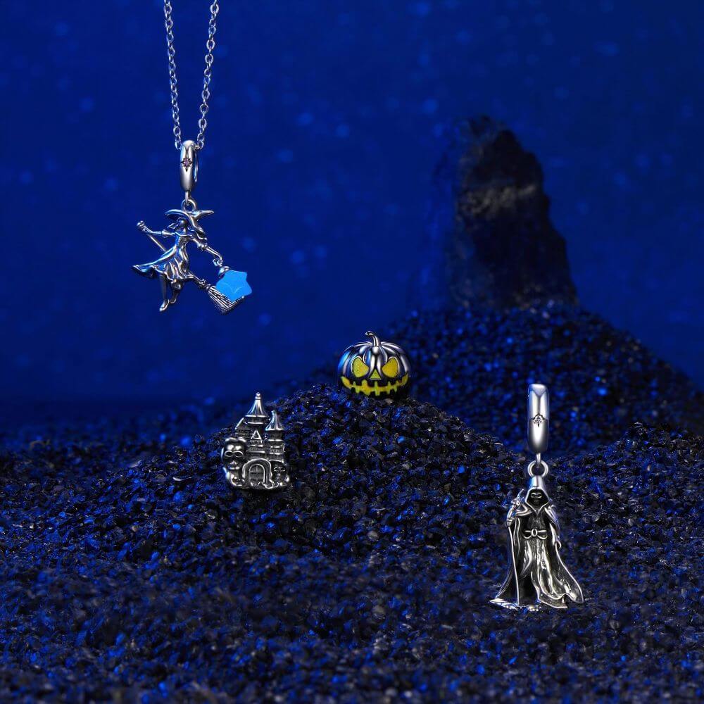 A set of silver fantasy-themed jewelry charms displayed on dark blue textured sand. The collection includes a silver witch on a broomstick pendant with a blue enamel accent, a silver castle charm, a silver pumpkin charm with yellow enamel, and a silver hooded figure charm.